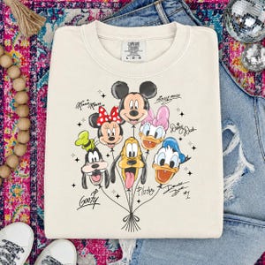 May include: Cream-colored sweatshirt featuring a graphic of Disney characters as balloons, including Mickey Mouse, Minnie Mouse, Donald Duck, and Goofy. The characters' names are written in script. The sweatshirt has a Comfort Colors tag.
