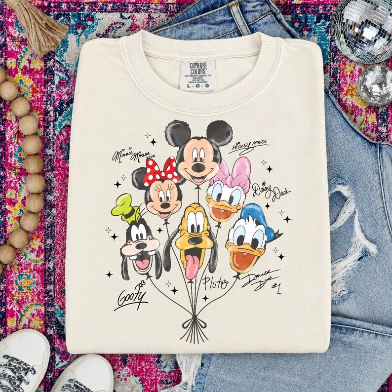 May include: Cream-colored sweatshirt featuring a graphic of Disney characters as balloons, including Mickey Mouse, Minnie Mouse, Donald Duck, and Goofy. The characters' names are written in script. The sweatshirt has a Comfort Colors tag.