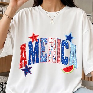 May include: White t-shirt with the word "AMERICA" in red, white, and blue patchwork letters. The design includes star and watermelon graphics, celebrating a patriotic theme.