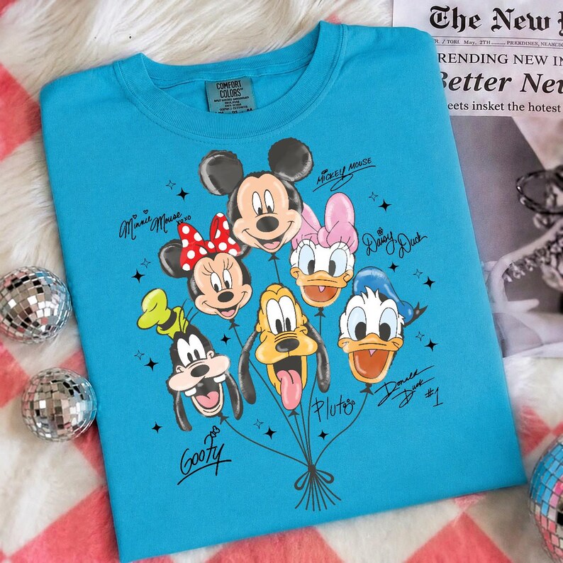May include: A turquoise Comfort Colors crew neck sweatshirt featuring a graphic of Mickey Mouse, Minnie Mouse, Daisy Duck, Donald Duck, Goofy, and Pluto as balloons. Each character's name is written in a script font. The shirt has a classic fit.