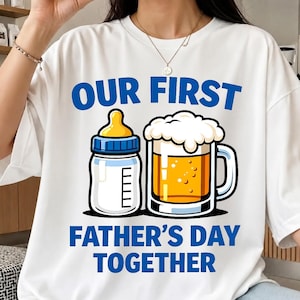 May include: White t-shirt with a blue and yellow graphic of a baby bottle and a beer mug. The text reads "OUR FIRST FATHER'S DAY TOGETHER" in blue block letters.