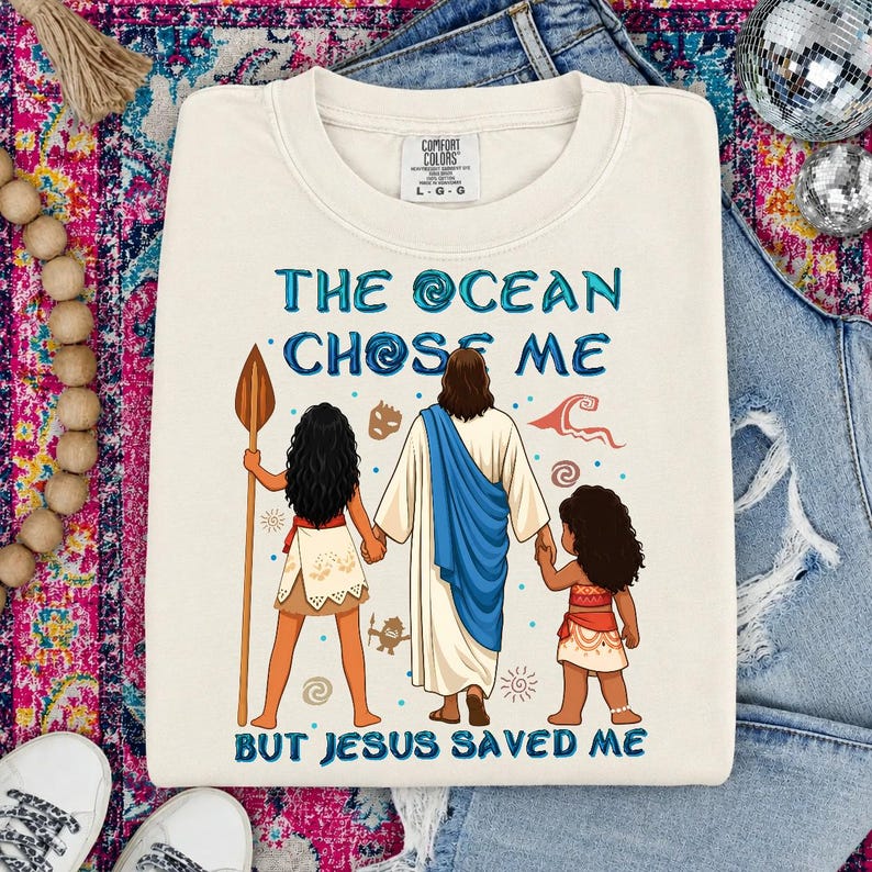 May include: Cream-colored Comfort Colors sweatshirt with the text "THE OCEAN CHOSE ME BUT JESUS SAVED ME" in blue. The graphic depicts three figures, including Jesus, and two girls. The sweatshirt is a crew neck and has long sleeves.