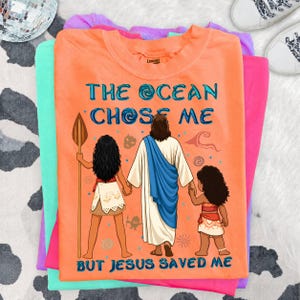 May include: Orange t-shirt with the text "THE OCEAN CHOSE ME BUT JESUS SAVED ME" and a graphic of Jesus holding hands with two girls. The shirt is part of a stack of other colorful t-shirts.