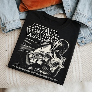 Op de afbeelding: Zwart Star Wars T-shirt met The Mandalorian en Grogu. Het shirt heeft het Star Wars logo in wit en de quote: "I'M THE MANDALORIAN. WEAPONS ARE PART OF MY RELIGION." Het shirt is op een licht oppervlak gevouwen.