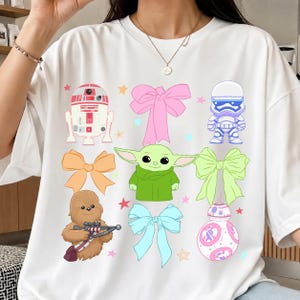 Op de afbeelding: Wit T-shirt met kleurrijke cartoon illustraties van Star Wars personages en strikken. Het ontwerp bevat R2-D2, een Stormtrooper, Chewbacca, Baby Yoda en BB-8, samen met roze, oranje, blauwe en groene strikken.