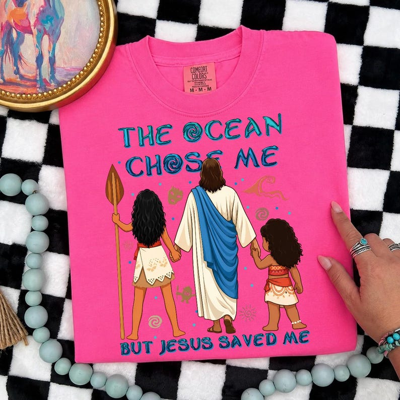 May include: A hot pink t-shirt with the text "THE OCEAN CHOSE ME BUT JESUS SAVED ME" in turquoise. The graphic depicts three figures, including Jesus, and two girls. The shirt is a Comfort Colors brand.