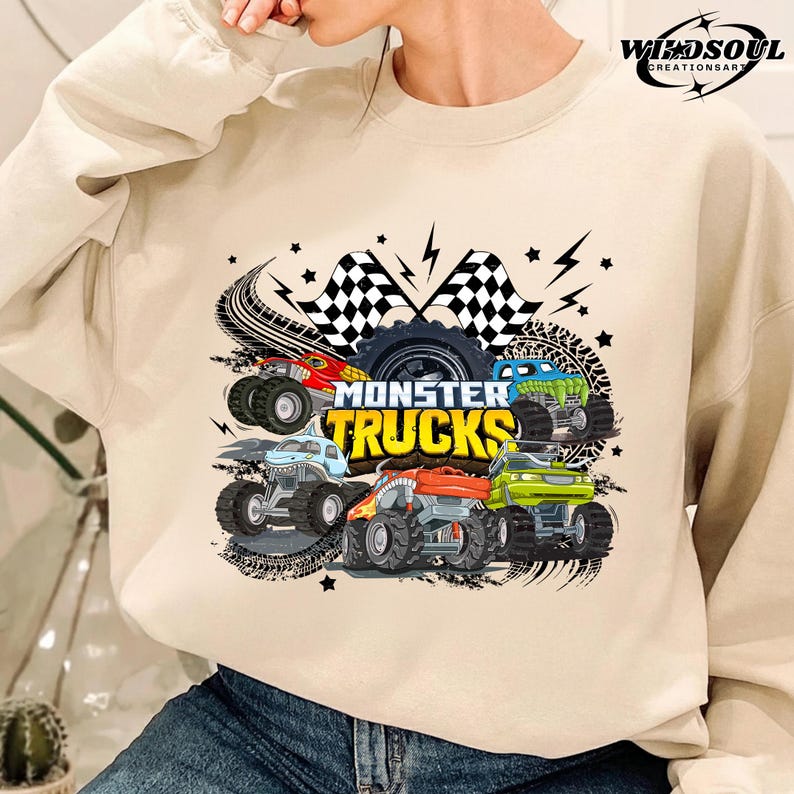 May include: Beige sweatshirt featuring a colorful graphic of monster trucks. The design includes five trucks in red, blue, green, orange, and light blue, with the words "Monster Trucks" in yellow. Black tire tracks, lightning bolts, and checkered flags complete the design.