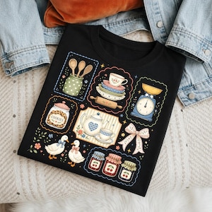 Pode incluir: T-shirt preto com um design temático de cozinha. O design apresenta ilustrações de chávenas, uma chaleira, uma balança, patos e frascos de geleia. As ilustrações são emolduradas com bordas decorativas e detalhes florais.