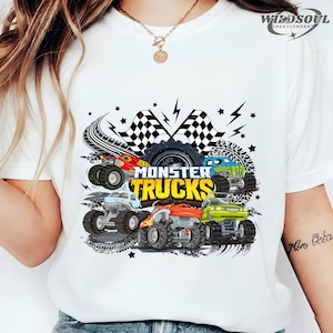 May include: White t-shirt featuring a colorful graphic of monster trucks. The design includes the text "MONSTER TRUCKS" in yellow, along with illustrations of various monster trucks in red, blue, green, and orange.