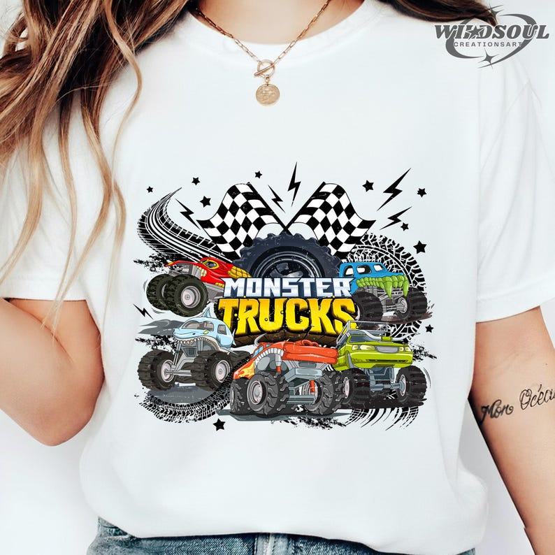 May include: White t-shirt featuring a colorful graphic of monster trucks. The design includes the text "MONSTER TRUCKS" in yellow, along with illustrations of various monster trucks in red, blue, green, and orange.