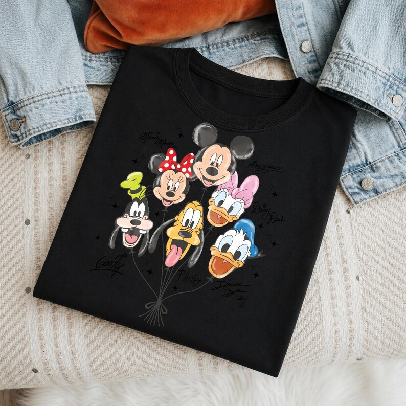May include: Black crew neck t-shirt featuring a graphic of Disney characters as balloons. The balloons include Mickey Mouse, Minnie Mouse, Goofy, Pluto, Daisy Duck, and Donald Duck. The characters' names are written on the shirt.