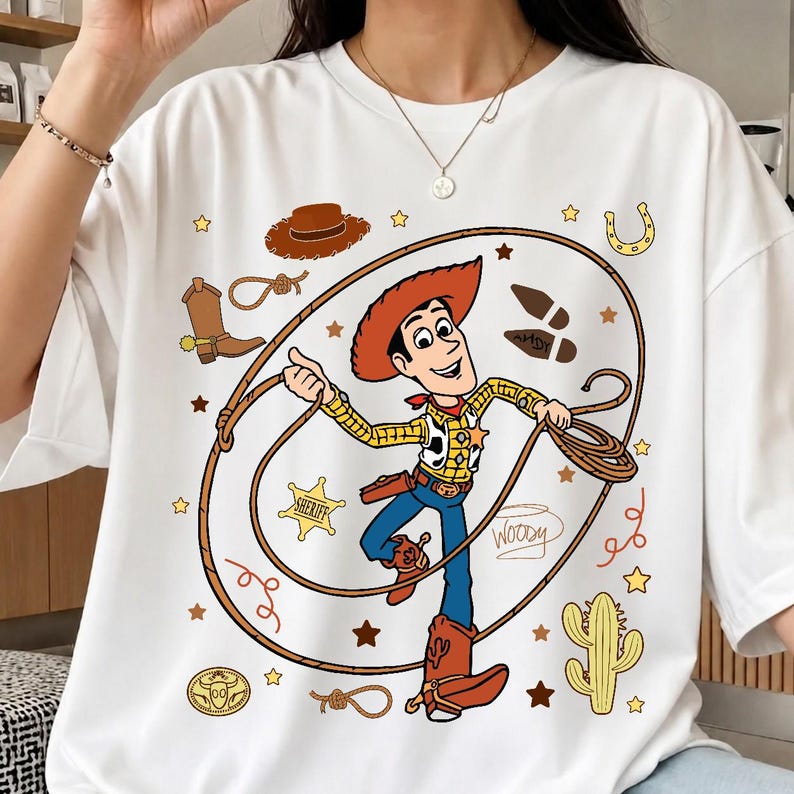 May include: A white t-shirt featuring a graphic print of the character Woody from Toy Story. The design includes Woody holding a lasso, surrounded by western-themed icons like a cowboy hat, boots, a sheriff badge, a cactus, and the name Woody.