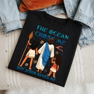 May include: Black t-shirt with the text "THE OCEAN CHOSE ME BUT JESUS SAVED ME" in blue. The shirt features a graphic of Jesus holding hands with two figures, one holding a spear. The figures are in a tropical setting.