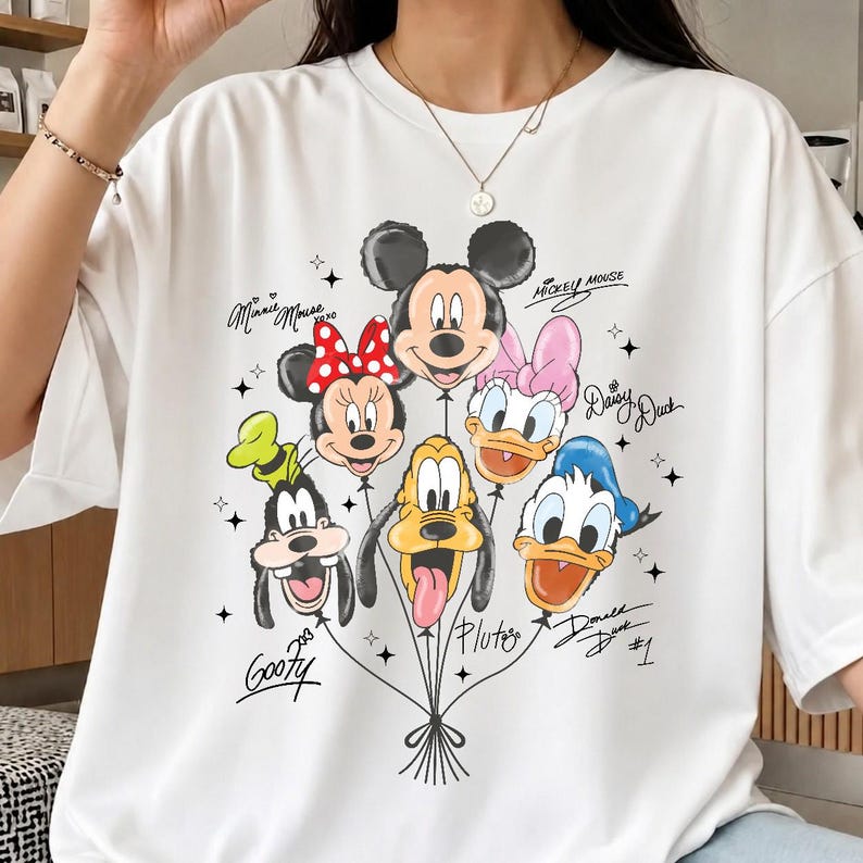 May include: White t-shirt featuring a colorful graphic of Disney characters as balloons. The design includes Mickey Mouse, Minnie Mouse, Donald Duck, Daisy Duck, Goofy, and Pluto. The shirt has a crew neck and short sleeves.