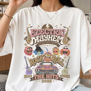 Könnte beinhalten: Weißes T-Shirt mit farbenfroher Grafik. Das Design zeigt den Text "ELECTRIC MAYHEM" und "ROCK ROLL", Cartoon-Figuren, Musikinstrumente und den Text "HEAR THE HITS SUMMER 2026".