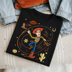 May include: A black t-shirt featuring a graphic of the character Woody from Toy Story, who is wearing a cowboy hat, yellow shirt, and blue pants. The design includes a lasso, a sheriff star, a cactus, a horseshoe, and the name Woody written in script.