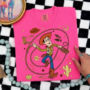 May include: A bright pink t-shirt featuring a graphic of the character Woody from Toy Story holding a lasso. The design includes a sheriff badge, a cowboy hat, a boot, a horseshoe, a cactus, and the name Andy written on a boot print.