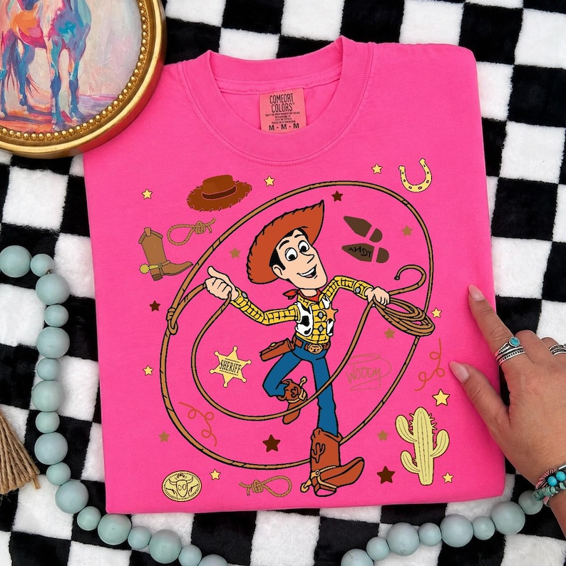 May include: A bright pink t-shirt featuring a graphic of the character Woody from Toy Story holding a lasso. The design includes a sheriff badge, a cowboy hat, a boot, a horseshoe, a cactus, and the name Andy written on a boot print.