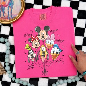 May include: A hot pink Comfort Colors t-shirt featuring a graphic of Mickey Mouse, Minnie Mouse, Donald Duck, Daisy Duck, Goofy, and Pluto as balloons. The characters' names are written in cursive. The shirt is medium size.