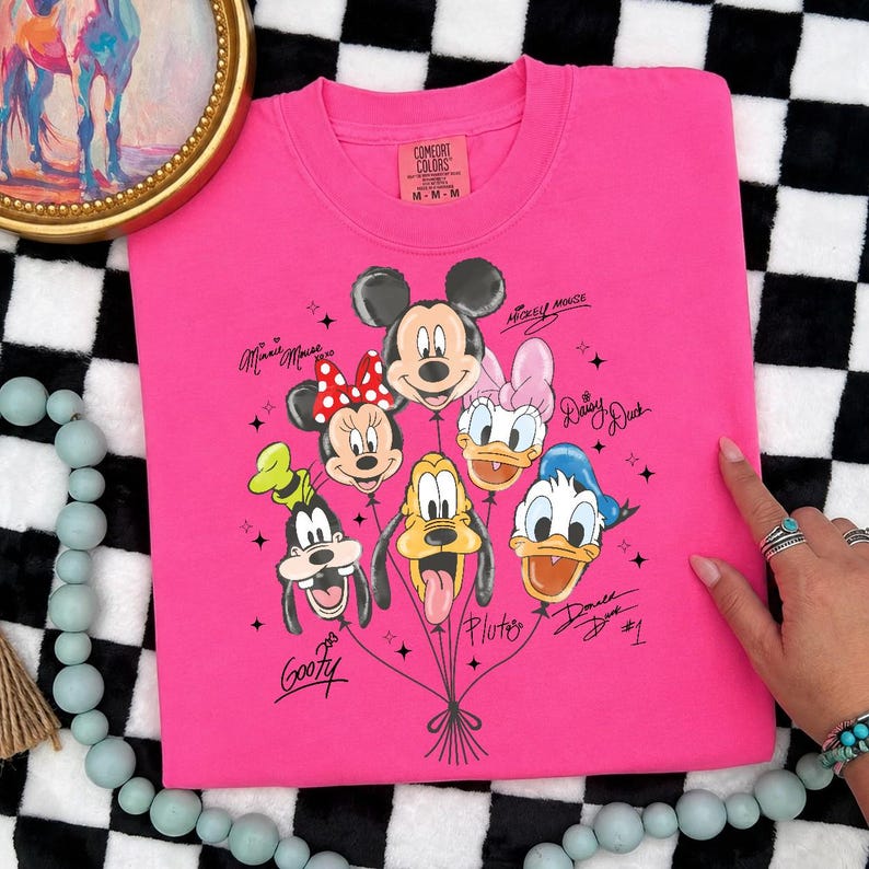 May include: A hot pink Comfort Colors t-shirt featuring a graphic of Mickey Mouse, Minnie Mouse, Donald Duck, Daisy Duck, Goofy, and Pluto as balloons. The characters' names are written in cursive. The shirt is medium size.
