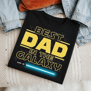 May include: Black t-shirt with the text "BEST DAD IN THE GALAXY" in yellow, with a blue lightsaber graphic. The shirt is folded on a light-colored surface, with a denim jacket visible in the background.