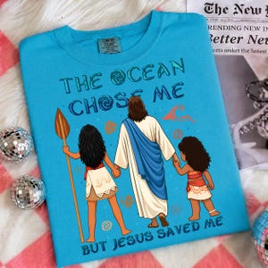 May include: A turquoise Comfort Colors t-shirt featuring the text "THE OCEAN CHOSE ME BUT JESUS SAVED ME" with an illustration of Jesus holding hands with two cartoon characters. The shirt has a crew neck.