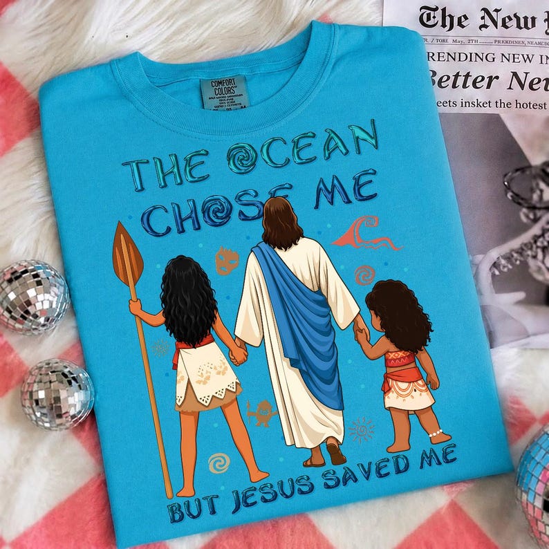 May include: A turquoise Comfort Colors t-shirt featuring the text "THE OCEAN CHOSE ME BUT JESUS SAVED ME" with an illustration of Jesus holding hands with two cartoon characters. The shirt has a crew neck.