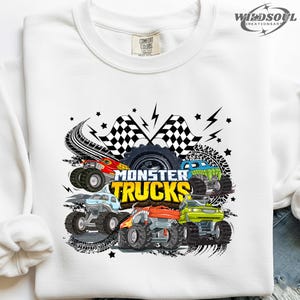 May include: White sweatshirt featuring a colorful graphic of monster trucks. The design includes multiple trucks in red, blue, green, and yellow, with the words "Monster Trucks" in yellow. Black tire tracks, stars, and checkered flags complete the design.