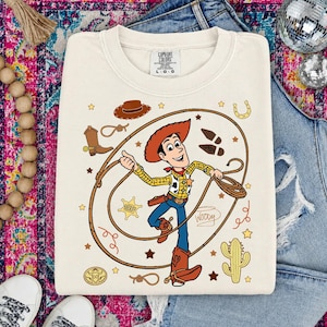 May include: A cream-colored crewneck sweatshirt featuring a graphic of the character Woody from Toy Story holding a lasso. The design includes a cowboy hat, a sheriff star, a cactus, a horseshoe, and the name Woody written in cursive script.