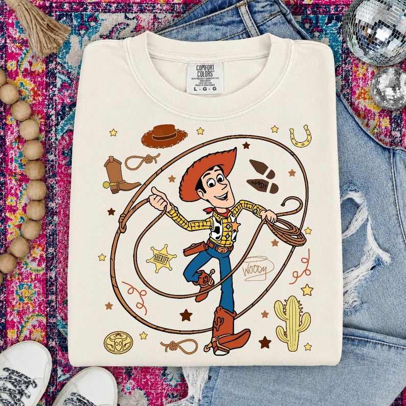 May include: A cream-colored crewneck sweatshirt featuring a graphic of the character Woody from Toy Story holding a lasso. The design includes a cowboy hat, a sheriff star, a cactus, a horseshoe, and the name Woody written in cursive script.
