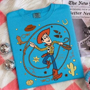 May include: A bright blue t-shirt featuring a graphic of the character Woody from Toy Story. The design includes a lasso, a sheriff badge, a cactus, a cowboy hat, a boot, and the name Andy written on a boot print, surrounded by small yellow stars.