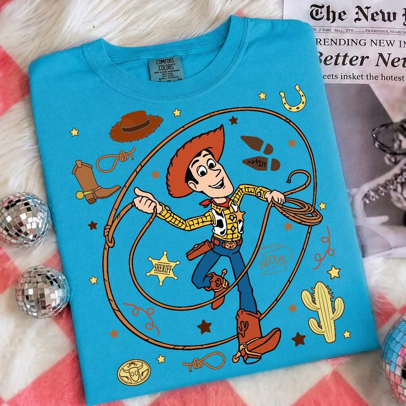 May include: A bright blue t-shirt featuring a graphic of the character Woody from Toy Story. The design includes a lasso, a sheriff badge, a cactus, a cowboy hat, a boot, and the name Andy written on a boot print, surrounded by small yellow stars.