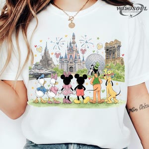 May include: White t-shirt featuring a colorful illustration of Disney characters and theme park landmarks. The design includes Mickey and Minnie Mouse, Donald and Daisy Duck, Goofy, and Pluto, with fireworks and castle imagery.