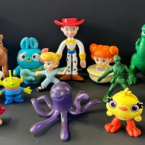 May include: A collection of plastic character figurines including a cowboy, a cowgirl, a green dinosaur, a brown horse, a potato-shaped figure, a blue rabbit, a green alien, a white spork character, a purple octopus, a yellow chick, and a soldier.