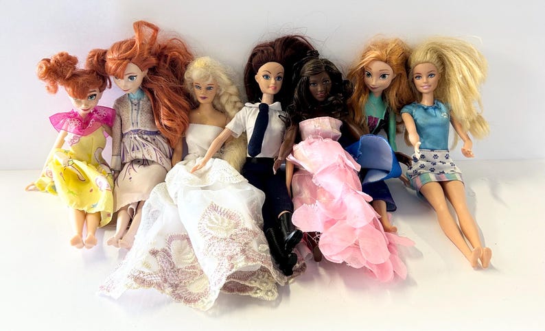 May include: A collection of fashion dolls with various hairstyles and outfits. The dolls include a variety of skin tones and wear dresses, skirts, and tops in colors like yellow, pink, blue, and white. Some dolls have long flowing hair, while others have their hair styled in buns.