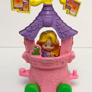 May include: A Little People toy featuring a pink tower with a purple roof and yellow accents. A blonde doll is inside the tower, with a green frog on the side. The toy has green wheels and two yellow signs with lantern graphics.