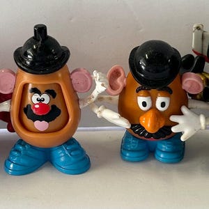May include: Four Mr. Potato Head toys in a row, each with different costumes. One is Captain America, another has a top hat, and the others have various outfits. The toys are on a white surface.