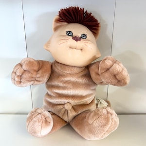 May include: A plush toy cat with a light brown body and a tan face. The cat has a tuft of brown hair on its head, blue eyes, and a small pink nose. The cat is wearing a matching light brown outfit.