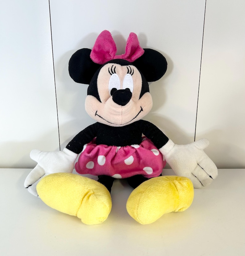 May include: A plush Minnie Mouse doll with a black body, pink skirt with white polka dots, and a pink bow. The doll has white gloves and yellow shoes. The doll is sitting upright.