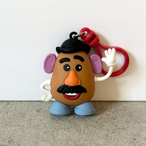May include: A brown potato-shaped character keychain featuring a black hat, purple ears, an orange nose, a black mustache, and blue shoes. The character has white arms and hands, with one hand raised in a wave, attached to a red plastic clip.