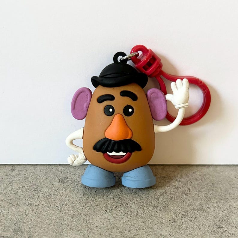 May include: A brown potato-shaped character keychain featuring a black hat, purple ears, an orange nose, a black mustache, and blue shoes. The character has white arms and hands, with one hand raised in a wave, attached to a red plastic clip.