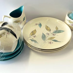 May include: A vintage cream-colored dinnerware set with a leaf and flower design in teal, yellow, and pink. The set includes plates, cups, saucers, a creamer, a sugar bowl, and a salt and pepper shaker.