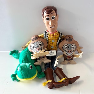 K&ouml;nnte beinhalten: Eine Sammlung von Artikeln zum Thema Toy Story, darunter eine Woody-Puppe, zwei Pl&uuml;schfiguren mit Schweinegesicht, ein gr&uuml;ner Pl&uuml;schfrosch und braune Cowboy-Stiefel mit gelben Sporen. Die Woody-Puppe tr&auml;gt ein gelbes kariertes Hemd und blaue Jeans.