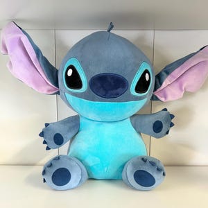 Może przedstawiać: Pluszowa zabawka postaci Stitch z filmu animowanego Lilo & Stitch. Zabawka jest gł&oacute;wnie niebieska z dużymi uszami, kt&oacute;re są r&oacute;żowe w środku. Ma duże czarne oczy i jasnoniebieski brzuch.
