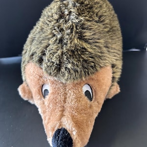 May include: A plush hedgehog toy featuring a soft, tan-coloured face with a black fabric nose and white-rimmed black eyes. The back of the toy is covered in textured, shaggy brown and tan faux fur that mimics the appearance of hedgehog quills.