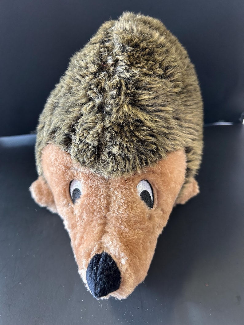 May include: A plush hedgehog toy featuring a soft, tan-coloured face with a black fabric nose and white-rimmed black eyes. The back of the toy is covered in textured, shaggy brown and tan faux fur that mimics the appearance of hedgehog quills.