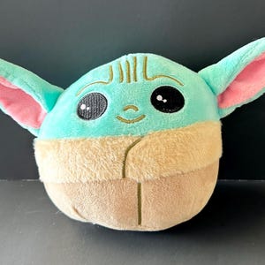 Pode incluir: Um brinquedo de pel&uacute;cia com um personagem com orelhas grandes, olhos grandes e pretos e um pequeno sorriso. O brinquedo tem uma cabe&ccedil;a e orelhas verde-claro com o interior das orelhas rosa. A parte inferior &eacute; castanha.