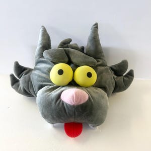 May include: A plush toy in the shape of a cartoonish monster head. The toy is primarily gray with large yellow eyes, a pink nose, and a red tongue sticking out. The toy has two pointed ears and two smaller ears.