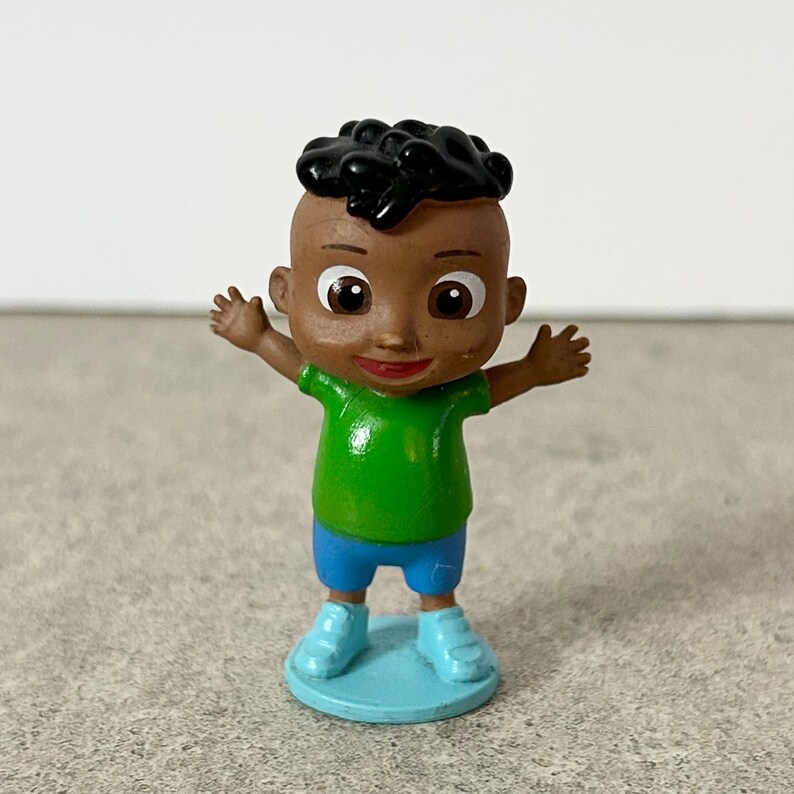 May include: A small figurine of a child with dark skin and black hair, wearing a bright green t-shirt and blue shorts. The character has large brown eyes, a small smile, and is standing on a light blue circular base with arms outstretched.
