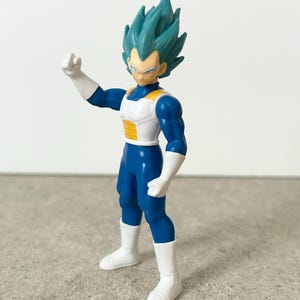 Pode incluir: Uma figura de a&ccedil;&atilde;o de pl&aacute;stico de um personagem com cabelo espetado azul-petr&oacute;leo, vestindo um traje azul com armadura de peito branca e amarela, luvas brancas e botas brancas, em uma pose com um bra&ccedil;o levantado e o punho fechado.
